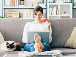 smiling woman sitting on sofa holding bank card looking at laptop