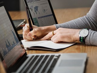person writing in notebook with two laptops open