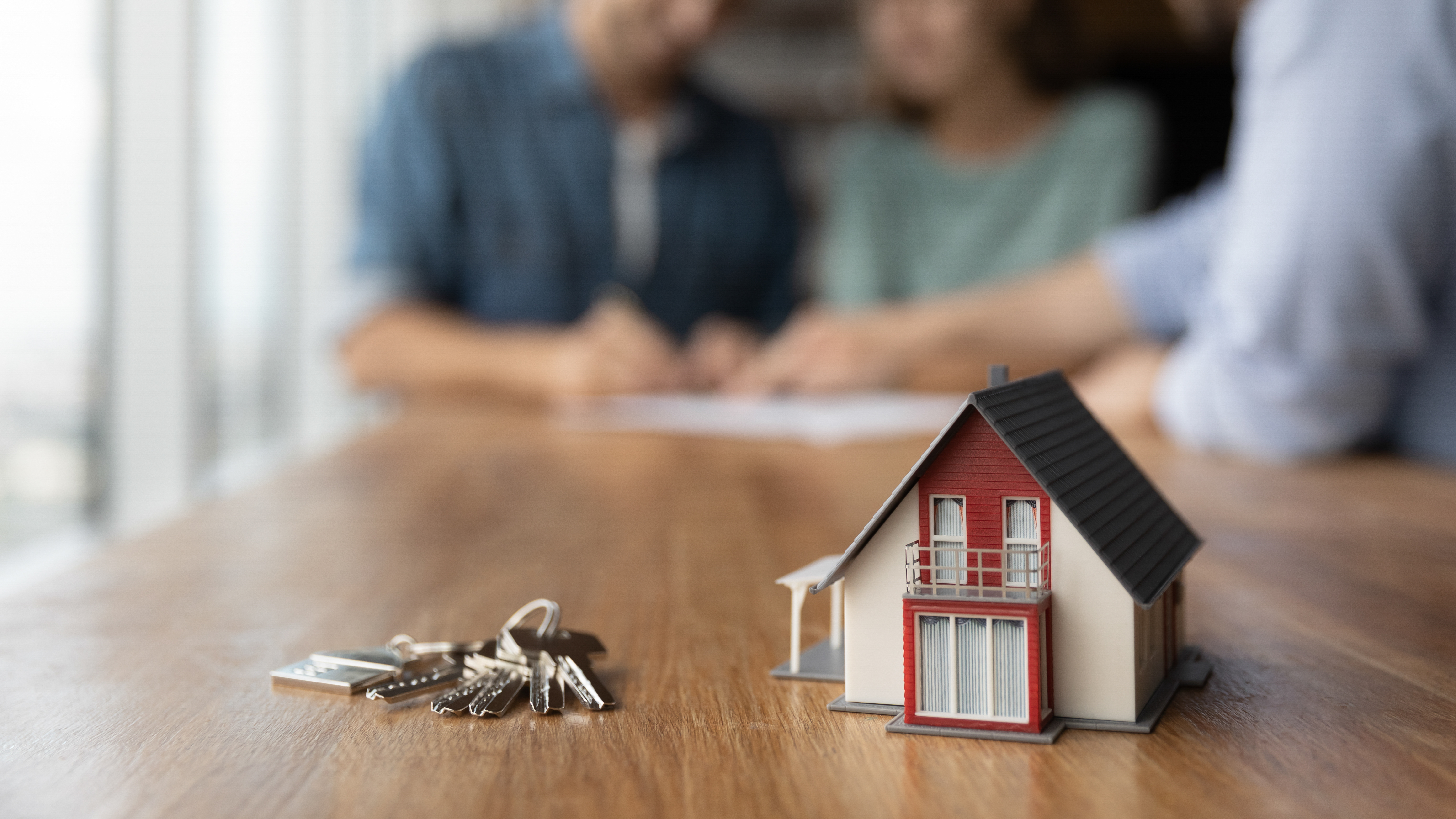 Foreground: Small toy house and keys on a table | Background: Couple signing mortgage paperwork