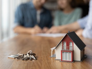 Foreground: Small toy house and keys on a table | Background: Couple signing mortgage paperwork